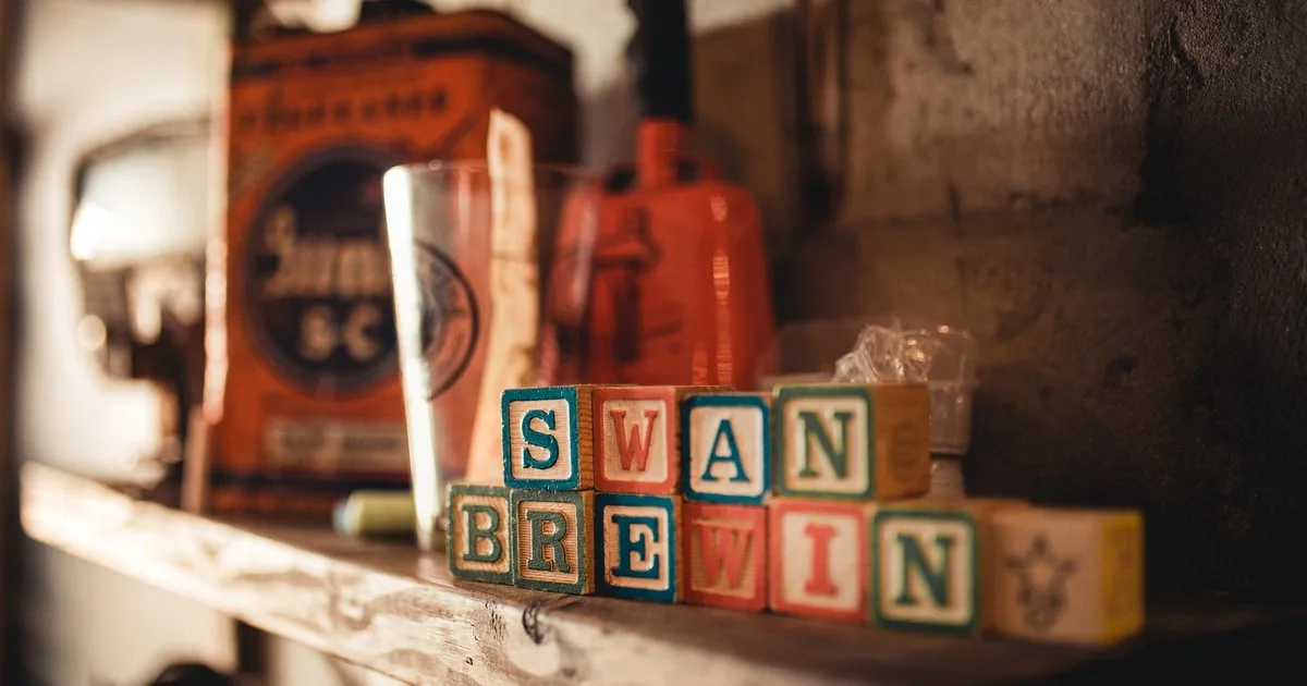 Vintage wooden letter blocks spelling SWAN BREWIN on a shelf at Swan Brewing, with an antique oil can and pint glass in the background
