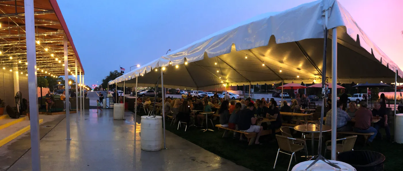 Packed covered patio at Swan Brewing at dusk — guests at picnic tables under a large white tent with string lights, pink and purple sunset sky behind