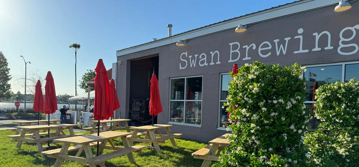 Swan Brewing exterior with picnic tables on the lawn and red umbrellas on a sunny day